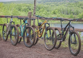 Four mountain bikes are lined up in a row on a dirt path next to a rustic, wooden fence. The background features a lush, green forest with a glimpse of a serene body of water, suggesting a natural and peaceful outdoor setting.