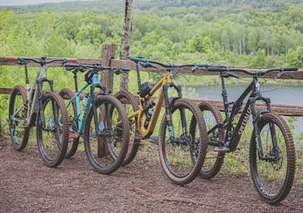 Four mountain bikes are lined up in a row on a dirt path next to a rustic, wooden fence. The background features a lush, green forest with a glimpse of a serene body of water, suggesting a natural and peaceful outdoor setting.