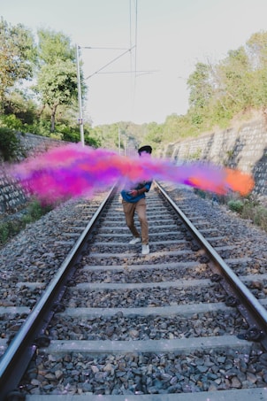 A person stands on train tracks surrounded by vibrant colored smoke. The tracks are lined with rocks and flanked by trees and greenery, with walls on either side. The smoke is a mix of pink, purple, and orange hues, creating a dramatic and artistic effect.