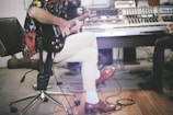 A person sitting on an office chair playing an electric guitar, with a colorful shirt and light pants, in a music studio. Nearby, another person is playing a keyboard on a desk with electronic equipment.