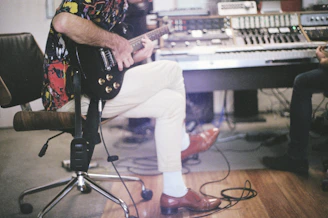 A person sitting on an office chair playing an electric guitar, with a colorful shirt and light pants, in a music studio. Nearby, another person is playing a keyboard on a desk with electronic equipment.