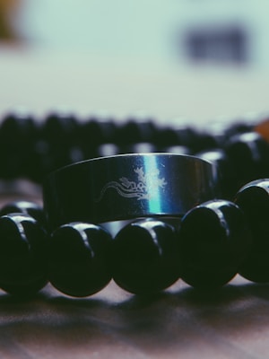 A metallic ring with an engraved design featuring a dragon motif is placed among glossy, black beads. The reflective surface of the ring captures light, creating a shiny effect, and the beads are arranged in a circular formation on a textured surface.