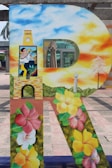 A large decorative letter 'R' is painted with vibrant imagery. The top section features a clear blue and orange sky with clouds, transitioning to a person in traditional attire, holding a musical instrument. Below, there is a floral design with hibiscus flowers in red, yellow, and pink. An umbrella and a small tree are also visible in the background.