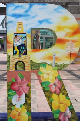 A large decorative letter 'R' is painted with vibrant imagery. The top section features a clear blue and orange sky with clouds, transitioning to a person in traditional attire, holding a musical instrument. Below, there is a floral design with hibiscus flowers in red, yellow, and pink. An umbrella and a small tree are also visible in the background.