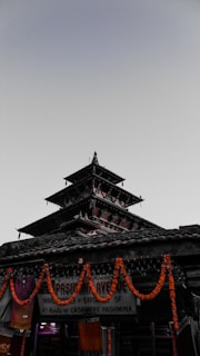 A traditional multi-tiered pagoda-style temple surrounded by garlands of orange marigold flowers. Below, there are signs for a store selling cashmere pashmina shawls. The sky is overcast, casting a dim light on the structure.