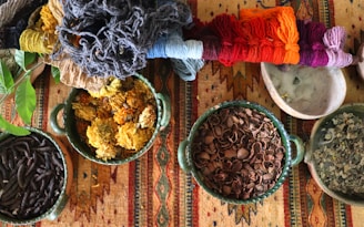 A collection of colorful yarns placed alongside bowls filled with various natural materials such as dried flowers, shells, and leaves. These items rest on a vibrant, patterned textile, showcasing a blend of earthy and bright hues.
