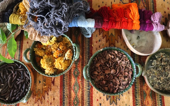 A collection of colorful yarns placed alongside bowls filled with various natural materials such as dried flowers, shells, and leaves. These items rest on a vibrant, patterned textile, showcasing a blend of earthy and bright hues.