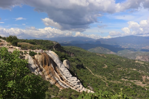 A scenic landscape featuring striking white geological formations that resemble waterfalls, set against a lush backdrop of green hills and expansive mountains under a partly cloudy sky. Small groups of people are visible walking near the edge of the formation, offering scale to the vastness of the panorama.