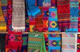A vibrant display of colorful, intricately woven textiles with a variety of geometric patterns. The fabrics feature a range of colors including reds, blues, greens, and yellows, and hang in layers, showcasing their diverse designs and textures.