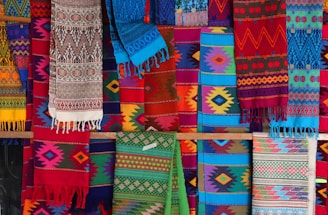A vibrant display of colorful, intricately woven textiles with a variety of geometric patterns. The fabrics feature a range of colors including reds, blues, greens, and yellows, and hang in layers, showcasing their diverse designs and textures.