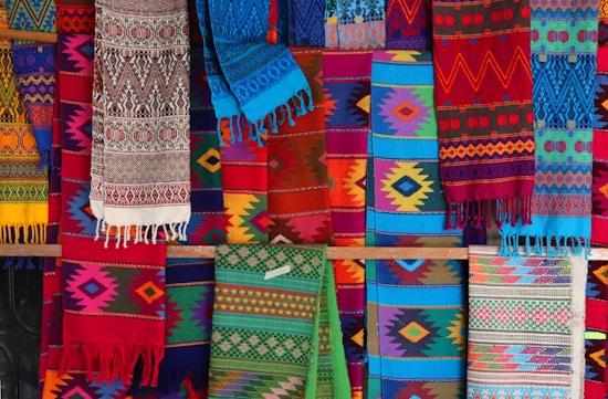 A vibrant display of colorful, intricately woven textiles with a variety of geometric patterns. The fabrics feature a range of colors including reds, blues, greens, and yellows, and hang in layers, showcasing their diverse designs and textures.