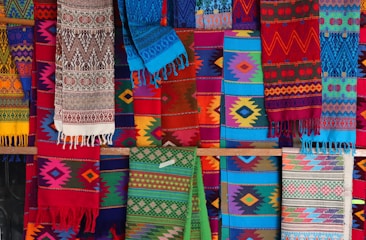 A vibrant display of colorful, intricately woven textiles with a variety of geometric patterns. The fabrics feature a range of colors including reds, blues, greens, and yellows, and hang in layers, showcasing their diverse designs and textures.