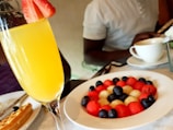 A brunch setting with a fresh mimosa garnished with a slice of strawberry. In the background, a colorful plate of assorted fruit balls including watermelon, melon, and blueberries. A person is seated at the table, enjoying a meal with a cup of coffee nearby.