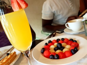 A brunch setting with a fresh mimosa garnished with a slice of strawberry. In the background, a colorful plate of assorted fruit balls including watermelon, melon, and blueberries. A person is seated at the table, enjoying a meal with a cup of coffee nearby.