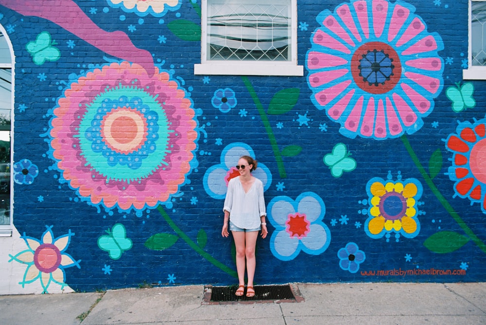 woman in grey dress shirt and blue distressed denim shorts standing in front of multicolored floral painted wall