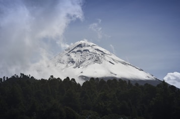 A majestic snow-capped mountain rises above a lush, green forest. Wisps of white clouds partially obscure the peak, giving a sense of mystery and grandeur. The sky is clear with a subtle gradient from light to darker blue.