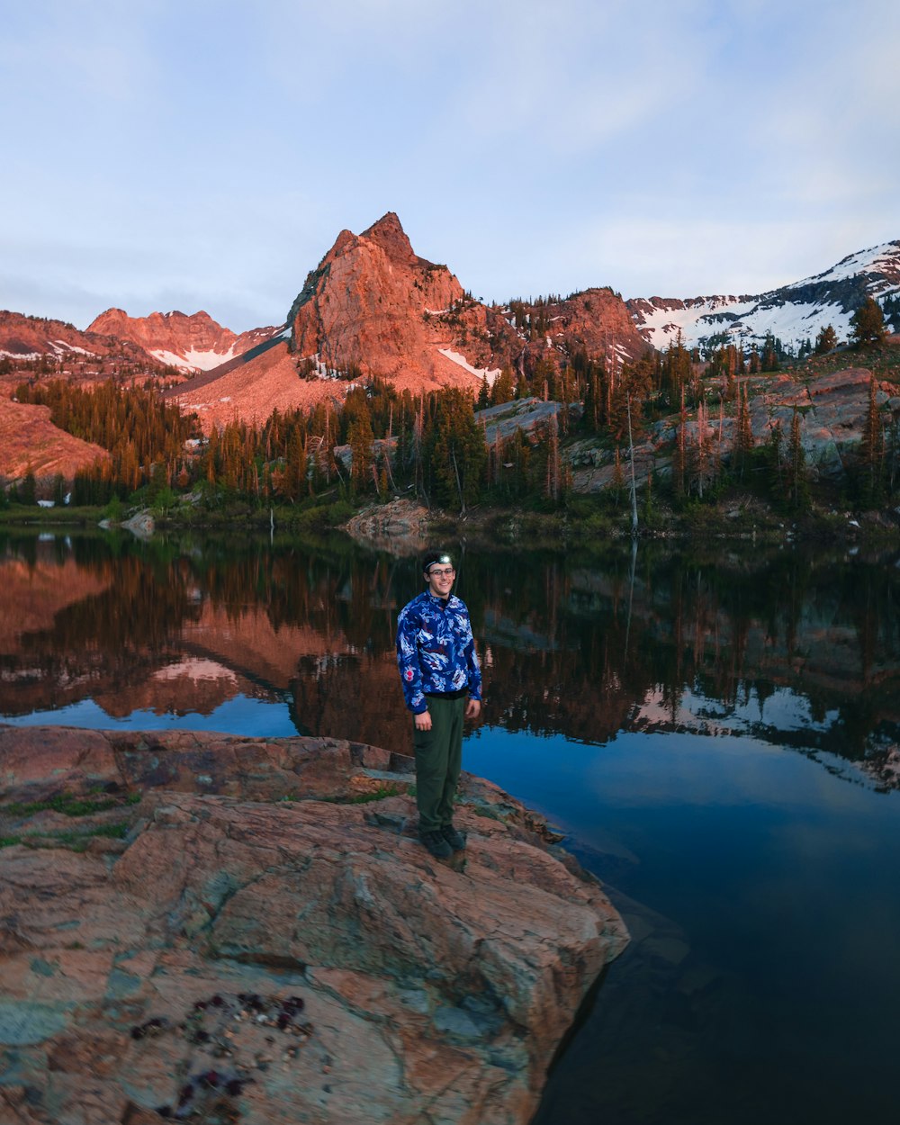 湖畔の茶色の岩の上に立つ青と白のジャケットと緑のズボンを着た男