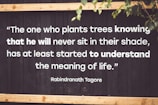 A motivational quote by Rabindranath Tagore is displayed on a dark wooden background with white, bold text. The quote emphasizes the virtue of selflessness and understanding the deeper meaning of life. Parts of green leaves are seen in the top right corner, adding a natural element.