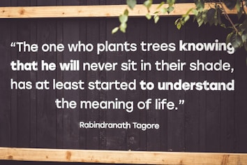 A motivational quote by Rabindranath Tagore is displayed on a dark wooden background with white, bold text. The quote emphasizes the virtue of selflessness and understanding the deeper meaning of life. Parts of green leaves are seen in the top right corner, adding a natural element.