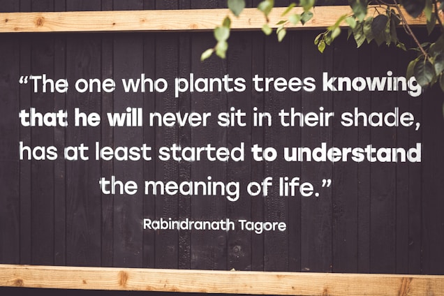 A motivational quote by Rabindranath Tagore is displayed on a dark wooden background with white, bold text. The quote emphasizes the virtue of selflessness and understanding the deeper meaning of life. Parts of green leaves are seen in the top right corner, adding a natural element.