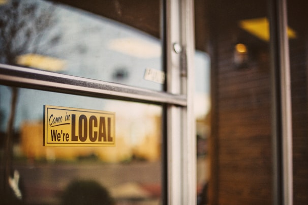 A sign on a glass door reads 'Come in We're Local', with reflections of a blurred outdoor scene in the glass. The background shows indistinct shapes and warm colors, suggesting an inviting and cozy environment.