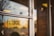 A sign on a glass door reads 'Come in We're Local', with reflections of a blurred outdoor scene in the glass. The background shows indistinct shapes and warm colors, suggesting an inviting and cozy environment.