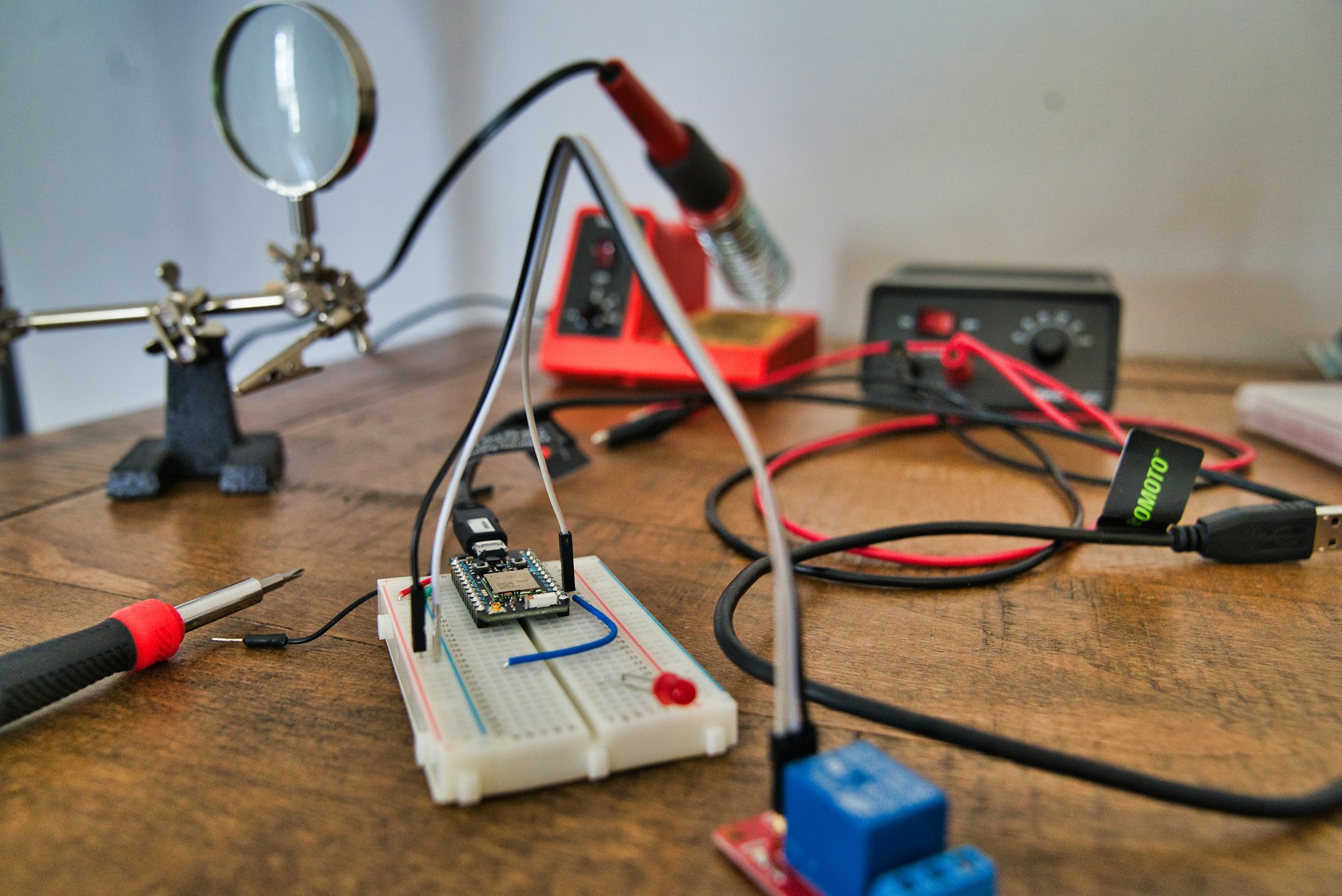 The Particle photon is an internet of things (IOT) device. In this picture it's hooked into a breadboard with an electrical relay attached to it. Also pictured in the background is a helping hand, a soldering iron, and a power supply.