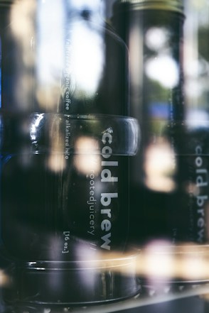 A close-up of bottles with labels reading 'cold brew' inside a refrigerator. The image has a slightly blurred, reflective surface, creating an artistic and moody look. The focus is on dark glass bottles, emphasizing the text and bottle shape amidst soft reflections.