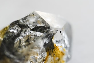 A close-up image of a raw, uncut gemstone or mineral with a rough texture and a multifaceted surface. The stone exhibits a mix of reflective and transparent qualities with metallic and earthy hues.