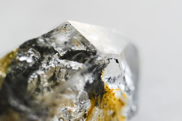 A close-up image of a raw, uncut gemstone or mineral with a rough texture and a multifaceted surface. The stone exhibits a mix of reflective and transparent qualities with metallic and earthy hues.