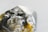 A close-up image of a raw, uncut gemstone or mineral with a rough texture and a multifaceted surface. The stone exhibits a mix of reflective and transparent qualities with metallic and earthy hues.