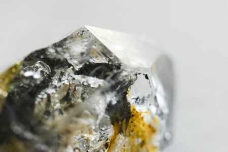 A close-up image of a raw, uncut gemstone or mineral with a rough texture and a multifaceted surface. The stone exhibits a mix of reflective and transparent qualities with metallic and earthy hues.