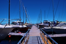 A marina with several sailboats and motorboats docked along narrow wooden piers. The sky is clear and blue, indicating a sunny day. Various boats display national flags, and the water is calm. Metal railings line the edges of the piers.