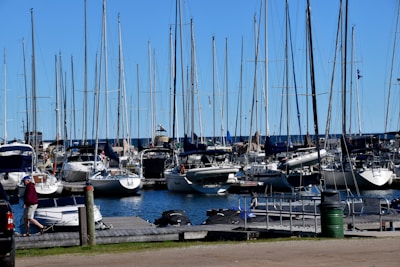 A marina filled with numerous sailboats docked side by side. The boats have tall masts and are aligned neatly. A blue sky spans overhead, and the water in the marina is calm. On the dock, a person wearing a red jacket and cap is walking past various equipment. In the foreground, a small motorboat is tied to the pier.