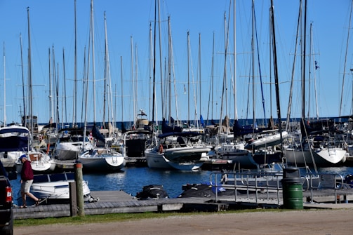 A marina filled with numerous sailboats docked side by side. The boats have tall masts and are aligned neatly. A blue sky spans overhead, and the water in the marina is calm. On the dock, a person wearing a red jacket and cap is walking past various equipment. In the foreground, a small motorboat is tied to the pier.