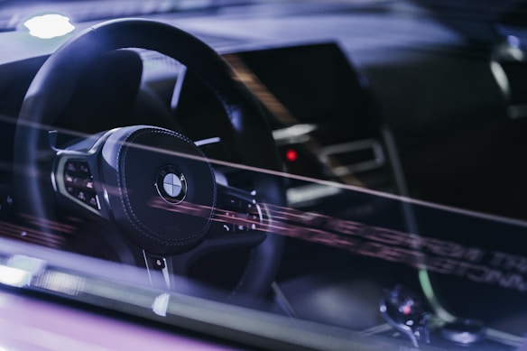 The image captures the interior of a car, focusing on the steering wheel with the BMW logo prominently displayed. Reflections can be seen on the windshield, adding a futuristic and sleek feel. The dashboard area is visible, though out of focus, suggesting a modern vehicle interior.