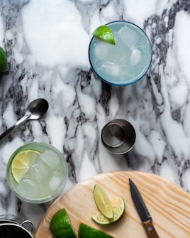 A marble surface displays a scene with two glasses filled with iced drinks garnished with lime wedges. A spoon, a metal jigger, and a cutting board with sliced limes are also visible. A knife is placed on the board next to the lime slices.