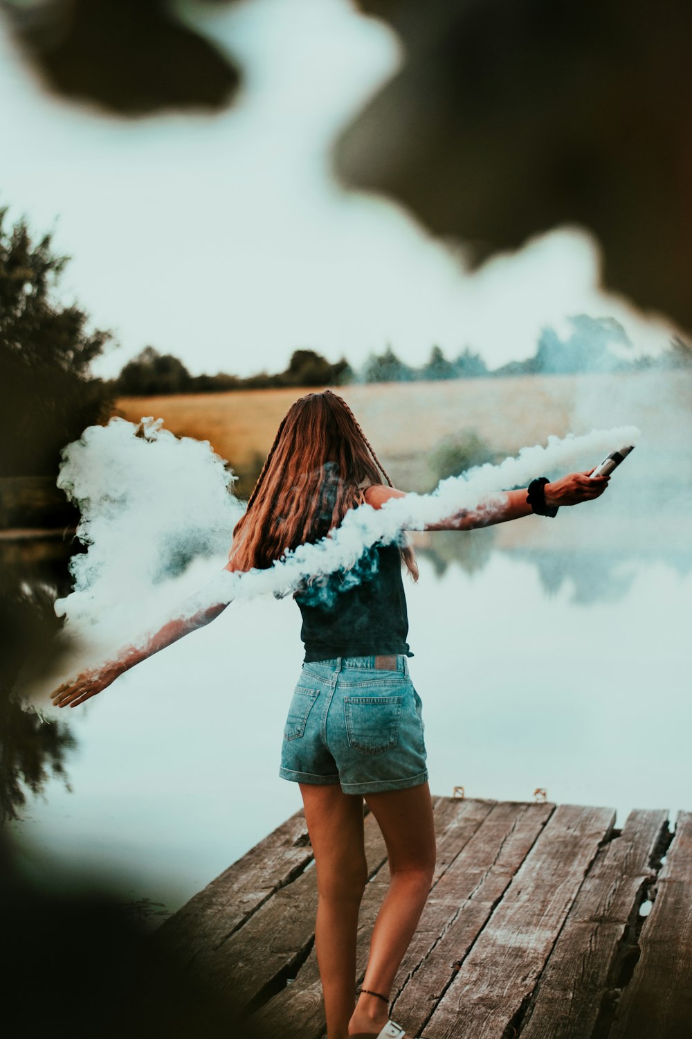 woman in blue denim shorts holding white smoke and standing on brown dock