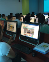 A group of people seated in a computer lab, each facing a desktop computer. The monitors display a website with text and colorful graphics. The room is dimly lit, with a presentation projected on a screen in the background. One person is holding a phone, and there are books or notebooks on the desk.