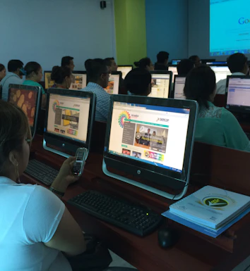 A group of people seated in a computer lab, each facing a desktop computer. The monitors display a website with text and colorful graphics. The room is dimly lit, with a presentation projected on a screen in the background. One person is holding a phone, and there are books or notebooks on the desk.
