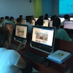 A group of people seated in a computer lab, each facing a desktop computer. The monitors display a website with text and colorful graphics. The room is dimly lit, with a presentation projected on a screen in the background. One person is holding a phone, and there are books or notebooks on the desk.