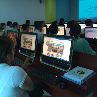 A group of people seated in a computer lab, each facing a desktop computer. The monitors display a website with text and colorful graphics. The room is dimly lit, with a presentation projected on a screen in the background. One person is holding a phone, and there are books or notebooks on the desk.