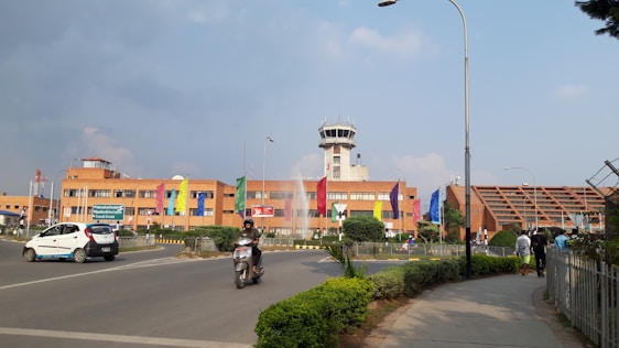 A large brick building with a central control tower, surrounded by various colored flags. There are vehicles on the road, including a car and a motorcycle. The sky is partly cloudy, and there is greenery along the path.