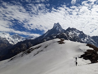 A snow-covered mountain landscape with a prominent peak under a sky filled with scattered clouds. Two people can be seen trekking along the snowy path, adding a sense of scale to the massive mountains in the background.