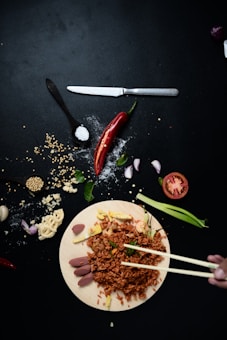 A dark background highlights a wooden board with a serving of fried rice garnished with vegetables. Surrounding the board are scattered ingredients like chopped red chilies, onions, garlic cloves, a knife, and a spoonful of salt. A hand holding chopsticks approaches the rice from the side.
