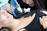 A tattoo artist in black gloves is focused on creating a tattoo on a client's upper chest area. The client, lying down with a slight smile, displays a sense of comfort. The tattoo machine is wrapped in a blue protective cover.