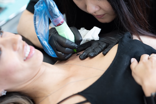 A tattoo artist in black gloves is focused on creating a tattoo on a client's upper chest area. The client, lying down with a slight smile, displays a sense of comfort. The tattoo machine is wrapped in a blue protective cover.