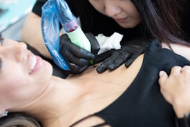 A tattoo artist in black gloves is focused on creating a tattoo on a client's upper chest area. The client, lying down with a slight smile, displays a sense of comfort. The tattoo machine is wrapped in a blue protective cover.