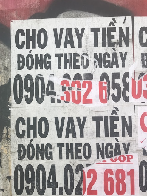 Several overlapping posters with bold text in Vietnamese advertising financial services. Some numbers are highlighted in red, indicating contact information. The posters are worn and layered on a rough urban surface.