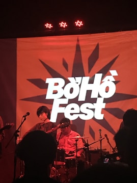 A band performs on stage with a large backdrop displaying the words 'Boho Fest' in bold lettering. The setting is dimly lit with red lighting, creating a warm atmosphere. There are musicians playing instruments such as a guitar and drums.